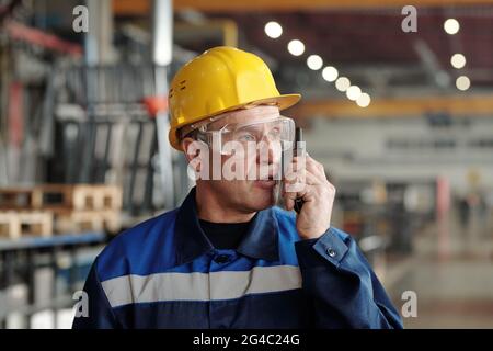 Cauccasico caucale sicuro e maturo in elmetto e occhiali di sicurezza che utilizzano un dispositivo radio per comunicare con i colleghi Foto Stock