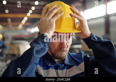 Uomo serio giovane sopportato in piedi alla fabbrica e mettendo il hardhat sulla testa mentre si prepara per il lavoro Foto Stock