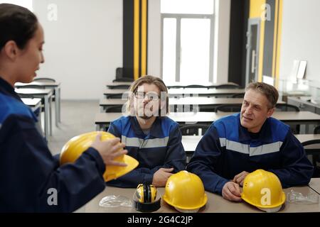 Lavoratori seduti a tavola con hardhats e occhiali in mensa di fabbrica e chiacchierando insieme Foto Stock