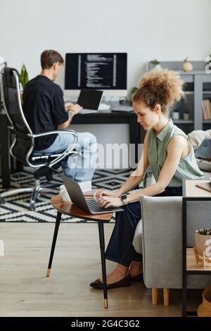 Donna d'affari matura seduta sul divano e che lavora su un computer portatile con il suo collega utilizzando il computer portatile anche in background Foto Stock