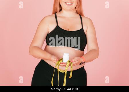 Primo piano sorridente più taglia giovane donna in outfit sportivo che tiene il nastro di misurazione e vaso di pillole di vitamina in piedi su sfondo rosa. Foto Stock