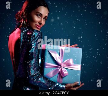 Giovane bella bruna donna positiva in abito nero scintillante in piedi indietro con regalo di festa in mani Foto Stock