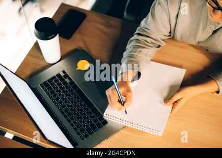 Giovane donna che studia corsi online con un computer portatile, scrivendo note in un notebook e bevendo caffè da una tazza riutilizzabile. Vista dall'alto, spazio per il testo Foto Stock