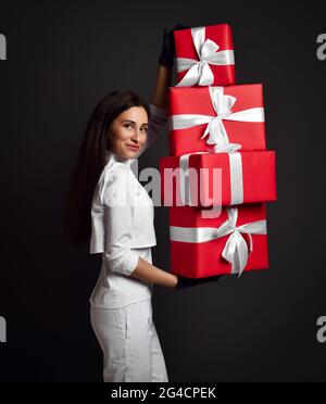 Attraente giovane positivo donna dentista medico in uniforme bianca e guanti in lattice in piedi e tenere un mucchio di scatole regalo di vacanza Foto Stock