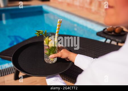 Una cameriera in camicia bianca serve un cocktail mojito per gli ospiti presso la piscina. Folle festa estiva all'aperto Foto Stock