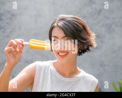 Donna che osserva i popsicles. Felice bella donna asiatica che indossa casual camicia bianca senza maniche che regge pop giallo, all'aperto. Sorridente femmina godendo Foto Stock