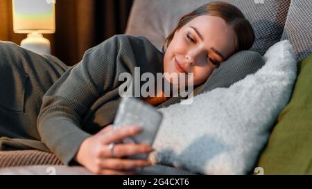 Felice ragazza sorridente che naviga su Internet o che chiacchiera con lo smartphone di notte sdraiata sul divano. La giovane donna nei vestiti domestici usa lo smartphone befor Foto Stock