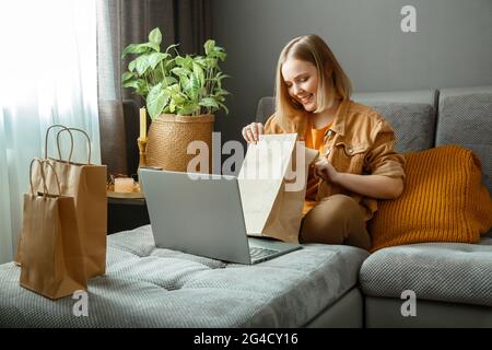 Felice giovane donna disimballare online ordini di merci o cibo. Acquisto online, consegna ordini. Adolescente ragazza rilassarsi sul divano considerando gli acquisti con Foto Stock