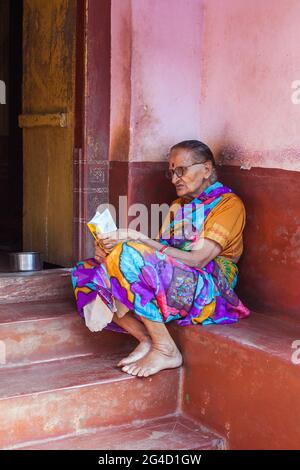 La donna indiana anziana si siede sul portico fuori del suo libro di lettura domestico, Gokarna, karnataka, India Foto Stock