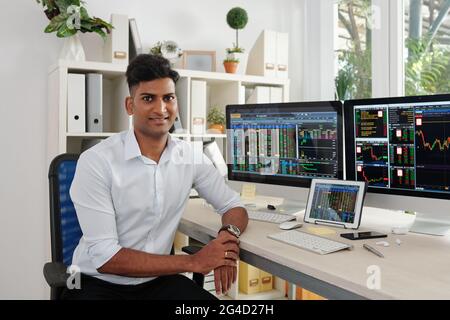 Ritratto di bel broker del mercato azionario seduto alla sua scrivania d'ufficio con computer con dati del mercato azionario in background Foto Stock