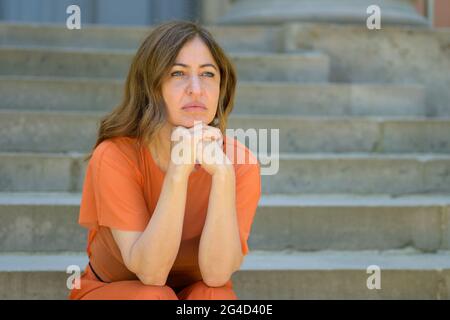 Annoiata donna seduta in attesa sui gradini all'aperto con espressione premurosa mentre appoggia il mento sulle sue mani guardando da parte Foto Stock