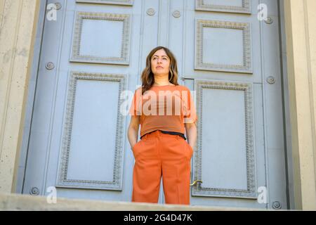 Vista ad angolo basso di una donna in colorato abito arancione in piedi in attesa con le mani in tasche di fronte a una grande storica porta di legno con copyspace Foto Stock