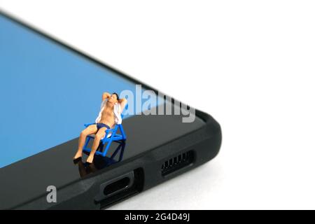 Foto di personaggi giocattolo in miniatura. Concetto di viaggio virtuale, sedile rilassante uomo sulla sedia sopra smartphone, isolato su sfondo bianco. Phot. Immagine Foto Stock