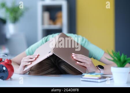 Stanca nello stress, la donna nasconde la testa con un computer portatile sul posto di lavoro. Foto Stock