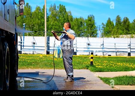 Autolavaggio per veicoli pesanti e cargo. Un uomo UN operaio del lavaggio dell'automobile in una maschera medica lava un camion con una pistola dell'acqua sulla strada in un giorno d'estate. Foto Stock