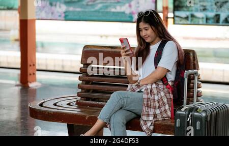 Donna asiatica in attesa di treno sulla stazione ferroviaria. Concetto di viaggio. Chi viaggia utilizzando uno smartphone per pianificare i viaggi online Foto Stock