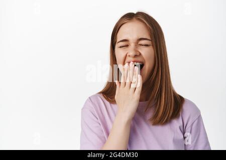 Primo piano di giovane donna che sbadisce, studentessa stanca durante gli esami, svegliandosi presto, coprendo la bocca aperta annoiata, in piedi esausta su sfondo bianco Foto Stock