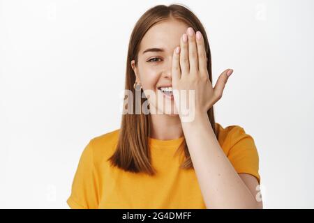 Primo piano ritratto di sorridente bionda ragazza, coprire metà del viso e ridere, guardando la fotocamera con un occhio, controllare la visione in negozio eyewear, in piedi Foto Stock