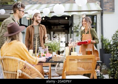 Gli amici si preparano per la cena di festa, servendo il tavolo con cibo delizioso nel bel cortile della casa di campagna Foto Stock