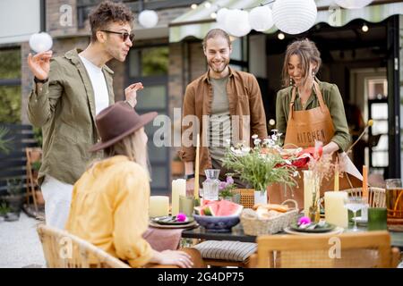 Gli amici si preparano per la cena di festa, servendo il tavolo con cibo delizioso nel bel cortile della casa di campagna Foto Stock