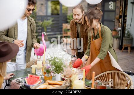 Gli amici si preparano per la cena di festa, servendo il tavolo con cibo delizioso nel bel cortile della casa di campagna Foto Stock