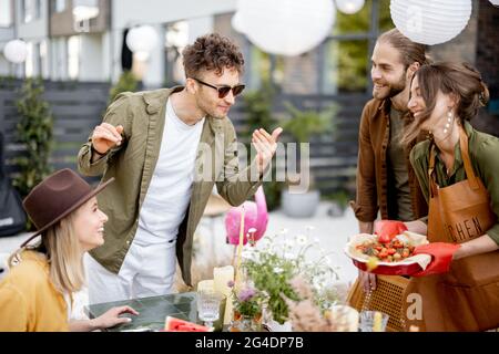 Gli amici si preparano per la cena di festa, servendo il tavolo con cibo delizioso nel bel cortile della casa di campagna Foto Stock
