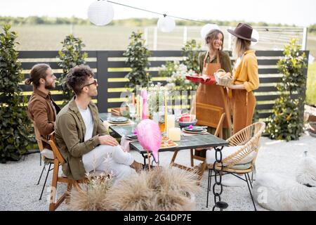 Gli amici si preparano per la cena di festa, due casalinghi che trasportano il pasto caldo e che servono il tavolo. Un gruppo di giovani che si riuniscono nel cortile della Foto Stock