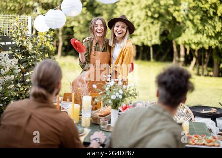 Amici che si preparano per la cena di festa, ritratto di due fidanzate in grembiule in piedi insieme. Un gruppo di giovani che si riuniscono nel cortile della Th Foto Stock
