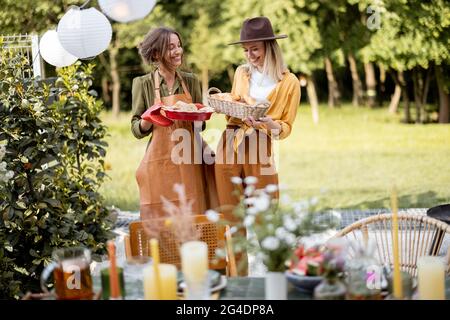 Gli amici si preparano per la cena di festa, due casalinghi che trasportano il pasto caldo e che servono il tavolo. Un gruppo di giovani che si riuniscono nel cortile della Foto Stock