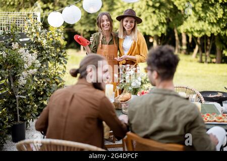 Amici che si preparano per la cena di festa, ritratto di due fidanzate in grembiule in piedi insieme. Un gruppo di giovani che si riuniscono nel cortile della Th Foto Stock