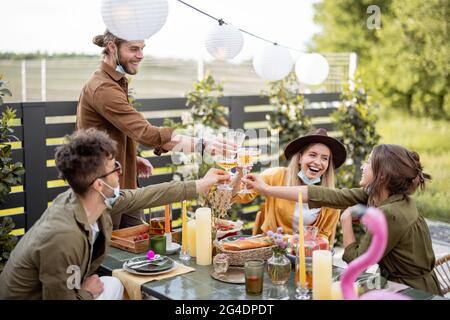 Giovani amici in maschera di faccia a cena festiva, occhiali da graffio sul cortile della casa di campagna. Felice di incontrarsi nella vita reale dopo p Foto Stock