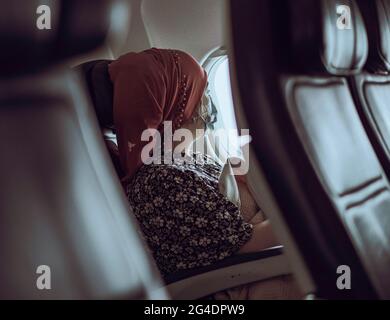 Una donna che dormiva viaggiatore si siede a una finestra di aeroplano. La donna che indossa la maschera si siede in una finestra soleggiata che dorme e guarda la vista fuori dalla finestra Foto Stock