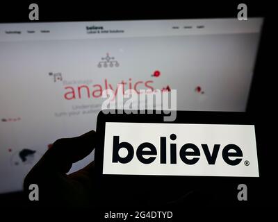 Persona che tiene il cellulare con il logo dell'etichetta francese del record Believe Digital sullo schermo di fronte alla pagina web aziendale. Mettere a fuoco il display del telefono. Foto Stock