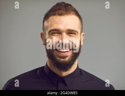 Primo piano ritratto di un uomo bearded in una camicia nera che sta sorridendo sinceramente su uno sfondo grigio. Foto Stock