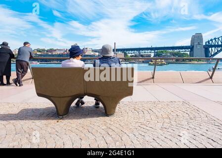 Due Signore asiatiche più anziane si siedono insieme su un posto a sedere all'aperto che guarda dalla Sydney Opera House fino al Sydney Harbour Bridge in Australia Foto Stock