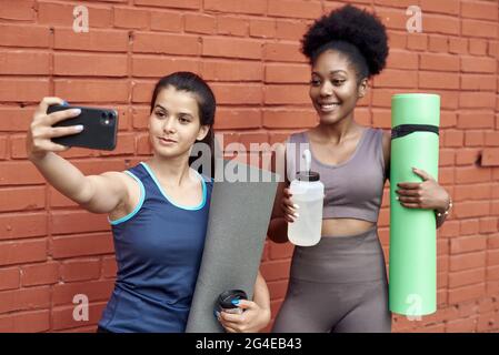 Immagine di donne atletiche giovani sorprendenti che prendono un selfie contro un muro di mattoni. Le donne nere sorridenti trascorrono il tempo insieme dopo l'allenamento sportivo. Foto Stock