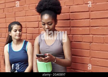 Immagine di donne atletiche giovani sorprendenti di fronte a un muro di mattoni, guardando nel telefono. Due amici di sesso femminile ridono al post sui social media. Foto Stock