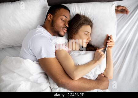 Giovane donna sdraiata sul letto tra le braccia del marito nero e utilizzando il telefono cellulare per scrivere il suo amante, vista dall'alto Foto Stock