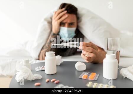 Uomo malato che mostra termometro con risultato di temperatura elevata alla macchina fotografica, sdraiato sotto coperta sul letto a casa Foto Stock