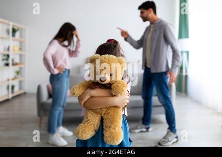 Concetto di conflitti familiari e bambini. La bambina coccolare l'orso del teddy in piedi contro i genitori che discutono Foto Stock