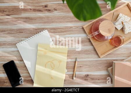 Blocco note con macchie di tè e vassoio con pentola e tazze sulla scrivania della persona creativa, vista dall'alto Foto Stock