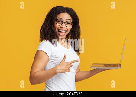 Concetto di persone e tecnologia. Gioiosa donna afroamericana che tiene il laptop e punta al gadget, sfondo giallo Foto Stock