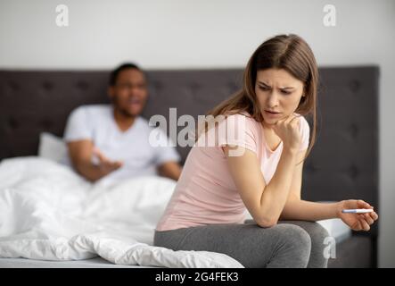 Infelice giovane donna seduta a letto con test di gravidanza, marito nero urlando a lei, al chiuso Foto Stock
