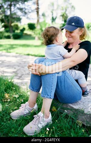 Il bambino si siede tra le braccia della madre, che si siede sul sentiero in pietra nel parco Foto Stock