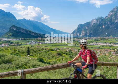 Bella donna anziana che cavalca la sua mountain bike elettrica negli uliveti di Arco e gode della vista sul Lago di Garda , Riva del Garda, Italia Foto Stock