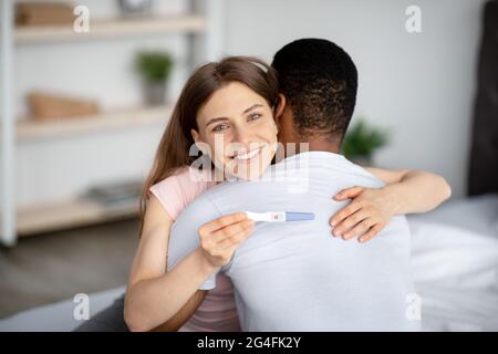 Sposato coppia multirazziale felice con il risultato positivo del test di gravidanza, abbracciandosi a casa Foto Stock