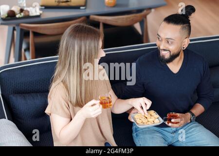 Sorridente giovane coppia bevendo tè con biscotti e discutendo notizie e gossip a casa Foto Stock