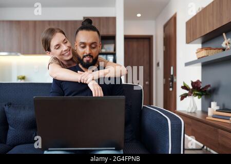 Felice giovane donna che agguenta il marito da dietro quando sta codificando sul laptop a casa Foto Stock