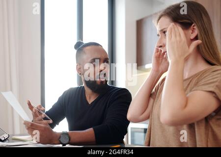 Scioccato uomo emotivo con fattura in mano guardando la moglie che ha dimenticato di pagare il pagamento mensile ipotecario Foto Stock