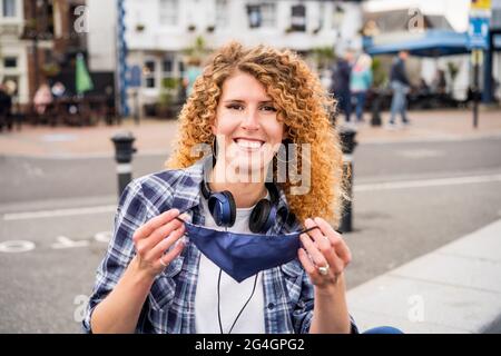 Giovane donna caucasica felice toglie una maschera protettiva che copre il viso all'aperto sullo sfondo della città europea. Pandemic Covid-19 è sopra concetto Foto Stock